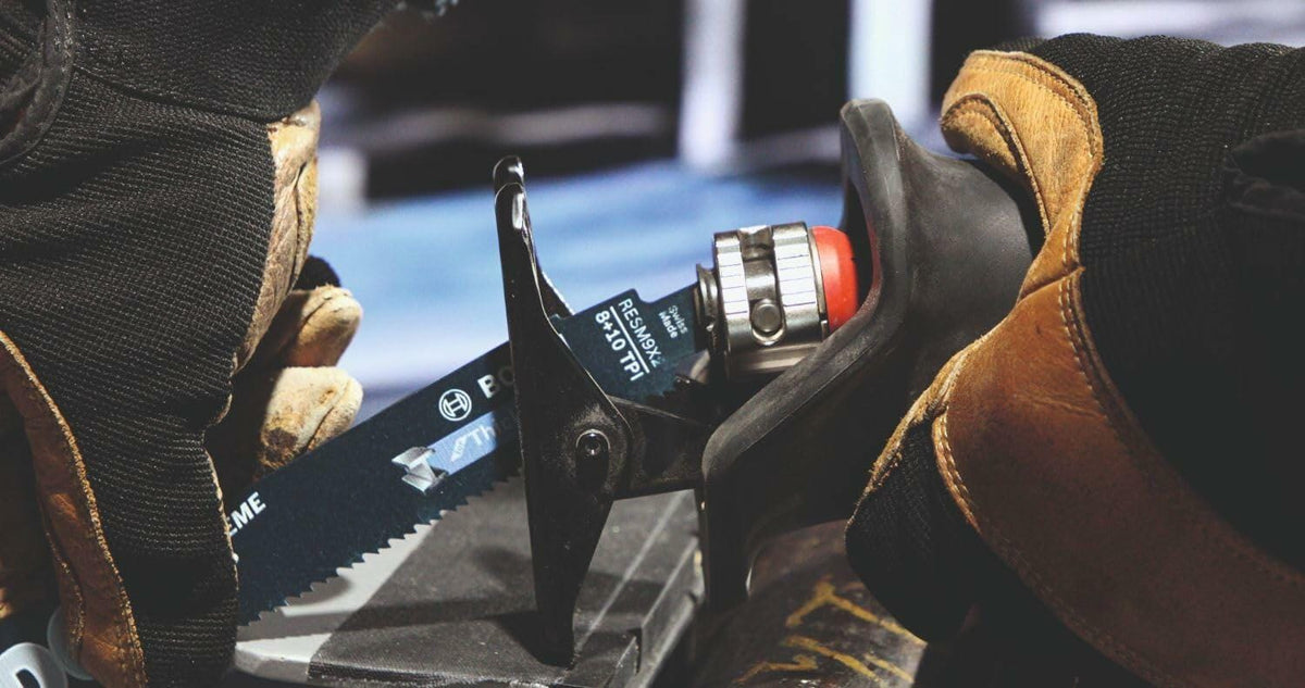 Close-up of gloved hands inserting a bi-metal reciprocating saw blade labeled “EXTREME” and “6 TPI” into a Bosch saw. Focus on blade clamp and safety handling in a construction setting.