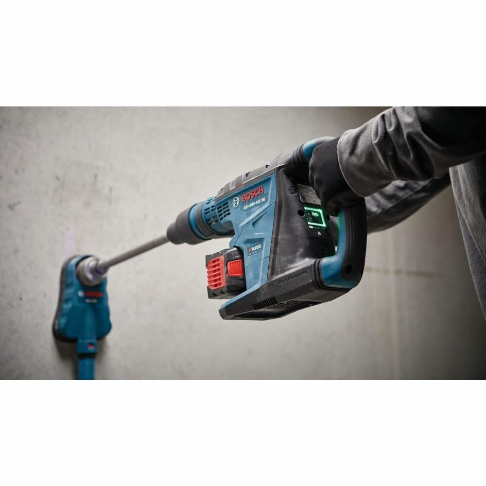 Person using Bosch rotary hammer drill with long bit to drill into concrete wall. Tool connected to dust extraction system. Worker wears gloves and long sleeves in a construction setting.