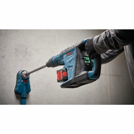 Person using Bosch rotary hammer drill with long bit to drill into concrete wall. Tool connected to dust extraction system. Worker wears gloves and long sleeves in a construction setting.