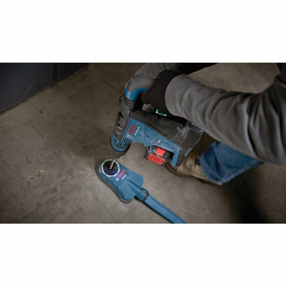 Person using Bosch rotary hammer drill with dust extraction attachment to drill into a concrete floor. Blue and black tool connected to a hose system. Gloves and construction attire suggest active jobsite use.