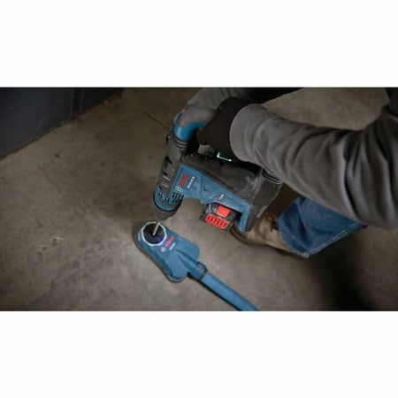 Person using Bosch rotary hammer drill with dust extraction attachment to drill into a concrete floor. Blue and black tool connected to a hose system. Gloves and construction attire suggest active jobsite use.