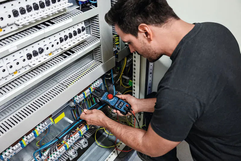 A technician running diagnostic maintenance inside a large commercial electrical equipment cabinet utilizing the Bosch PRO GFM1000-15 true RMS fork tester. The image showcases one handed functionality and high visibility reading capture on the high contrast inverted LCD display panel.
