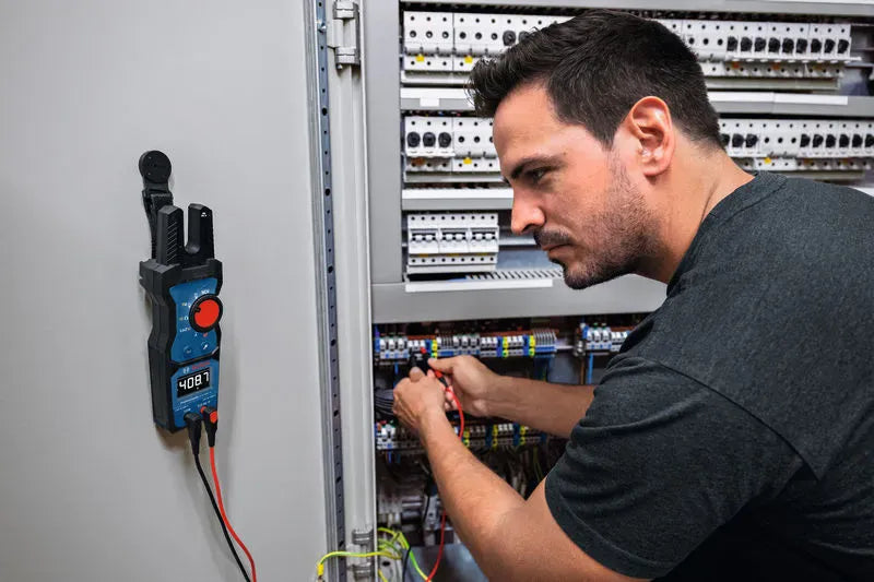 An industrial electrician troubleshooting a large commercial circuit panel while utilizing the Bosch PRO GFM1000-15 Open Fork Meter. The true RMS electrical tester is mounted hands free to the side of the metal cabinet door with a black magnetic hanging solution, clearly displaying a voltage reading on its high contrast inverted LCD panel.