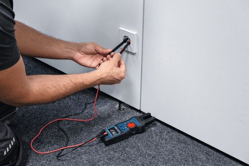 A close up view of a field technician inserting red and black premium silicone test leads into a wall outlet to run safety and voltage continuity tests. The Bosch PRO GFM1000-15 diagnostic electrical multi tester rests nearby on the carpeted floor, capturing real time system readings on its digital display panel.