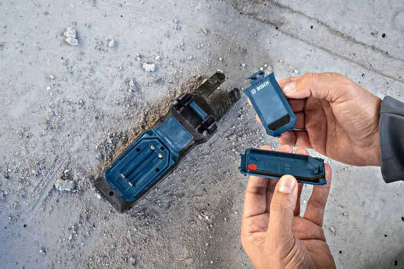 An overhead close up view of a technician's hands holding the unlatched battery bay door and sealing components away from the reverse side of the Bosch PRO GFM1000-15 Open Fork Meter. The image demonstrates the internal battery terminal layout and rugged jobsite engineering on a textured concrete surface.
