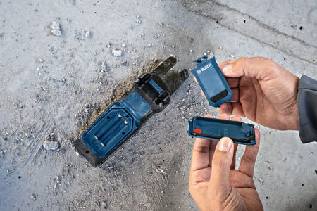 An overhead close up view of a technician's hands holding the unlatched battery bay door and sealing components away from the reverse side of the Bosch PRO GFM1000-15 Open Fork Meter. The image demonstrates the internal battery terminal layout and rugged jobsite engineering on a textured concrete surface.