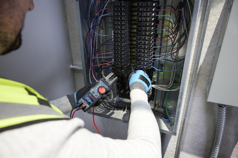 An electrician using the Bosch PRO GFM1000-15 Open Fork Meter to diagnose lines inside an open electrical panel breaker box. The technician uses the test leads and multi tester simultaneously to check residential or commercial electrical systems with safety certified CAT IV 600V protection.