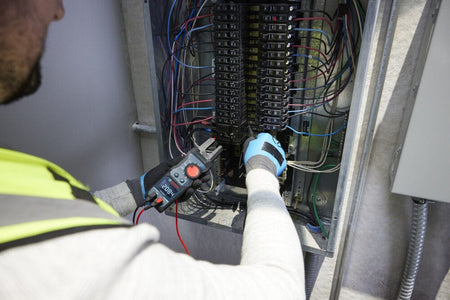An electrician using the Bosch PRO GFM1000-15 Open Fork Meter to diagnose lines inside an open electrical panel breaker box. The technician uses the test leads and multi tester simultaneously to check residential or commercial electrical systems with safety certified CAT IV 600V protection.