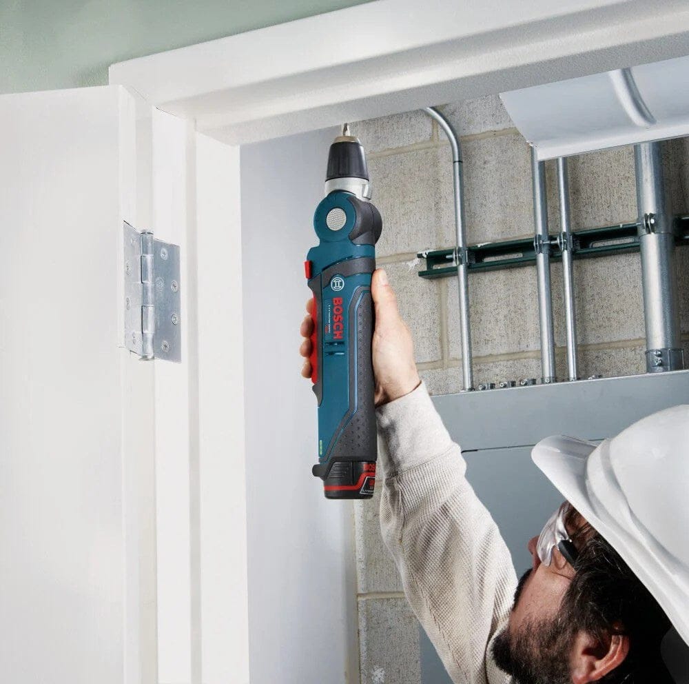 Action shot of a worker using the drill in a 180-degree straight configuration to drive a screw into the top of a white wooden door frame while working near an industrial electrical panel.