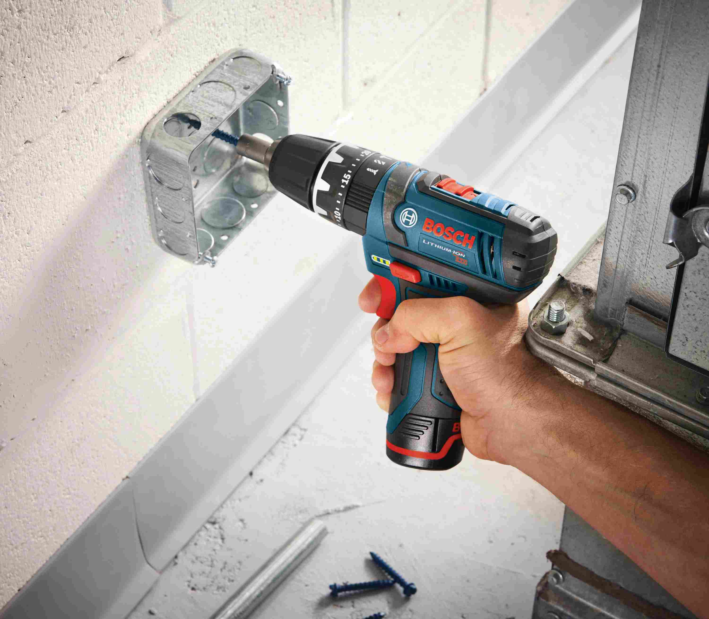A person holding the Bosch PS130-2A drill driver to fasten a screw inside a metal electrical junction box mounted on a white block wall.