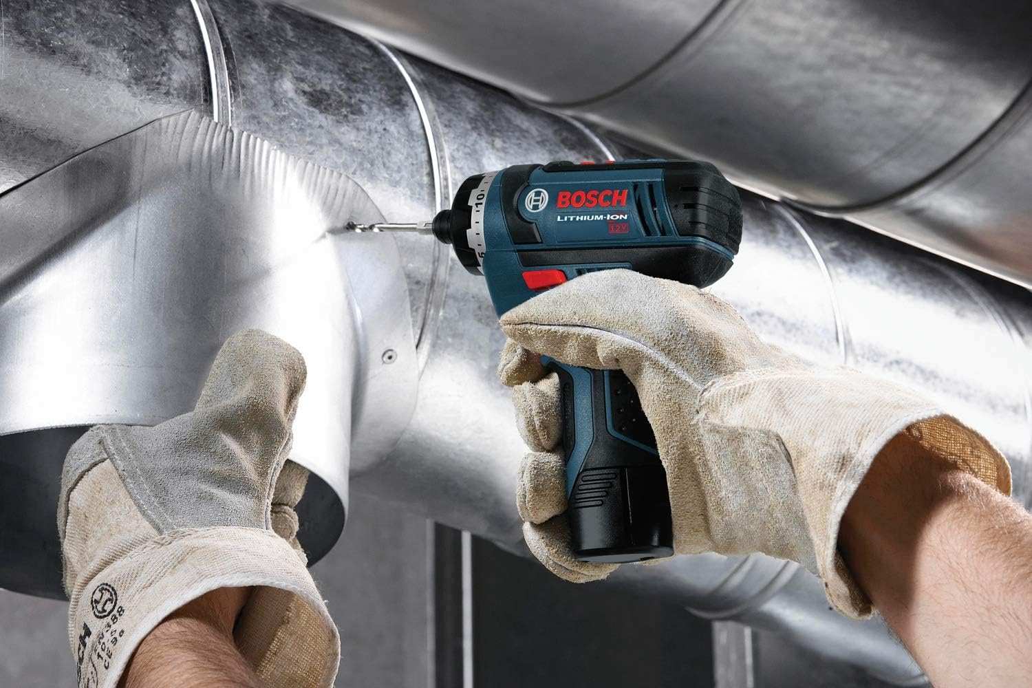 A professional using the compact Bosch 12V pocket driver to drill into galvanized metal ductwork in a tight, overhead industrial space.