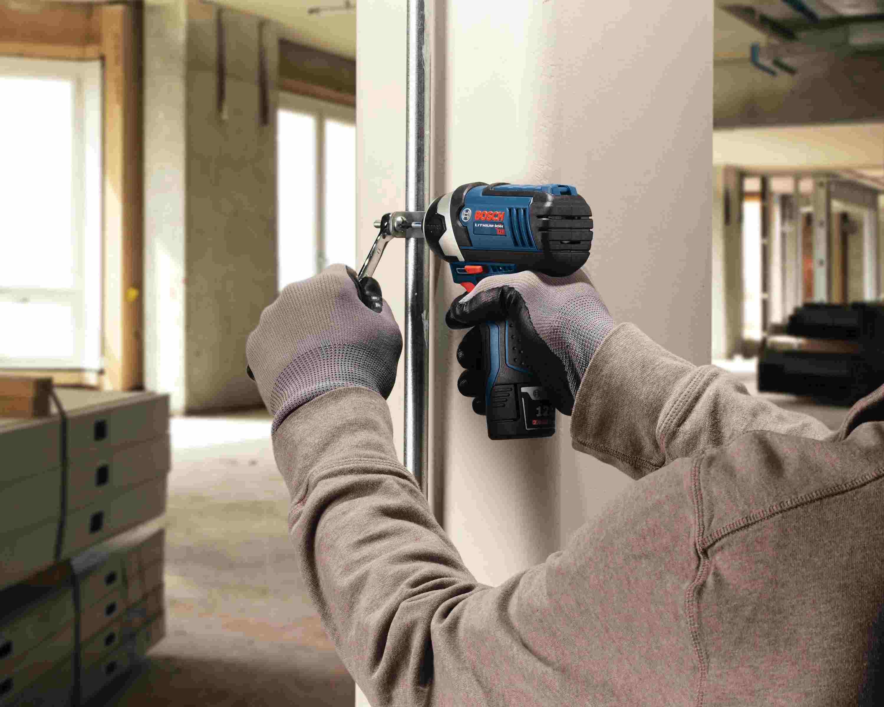 A construction worker wearing gloves using the Bosch PS41-2A impact driver with a socket adapter to tighten a bolt on a vertical metal pipe support while holding a wrench in the other hand.
