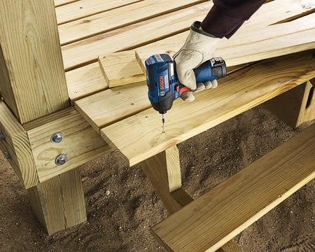 A professional contractor using the ultra-compact Bosch 12V Max brushless impact driver to drive a fastener into pressure-treated lumber on an outdoor construction project, demonstrating the tool's portability and power.
