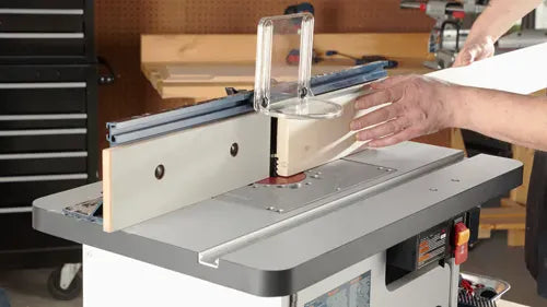 A perspective workshop photo showing a carpenter guiding a long piece of lumber stock across the laminated MDF router table top and against the tall aluminum fence with an adjustable clear safety guard.