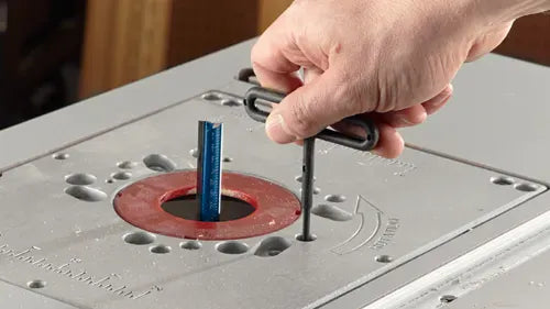 A close-up field photograph showing a woodworker using a black t-handle hex wrench to perform above-table router height adjustments through the precision aluminum mounting plate insert.