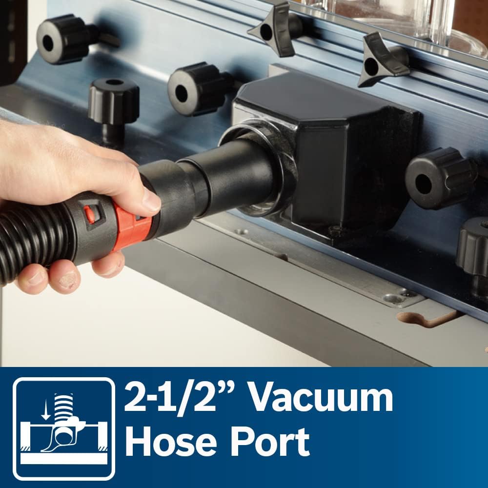 A detailed close-up product image showcasing a hand connecting a black vacuum hose with a red locking collar to the 2-1/2 inch dust collection port on the back of the aluminum router fence, with a lower blue banner reading 2-1/2 Inch Vacuum Hose Port.