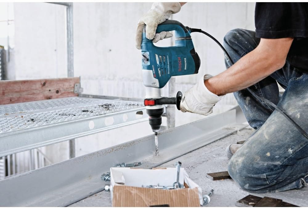 A real-world construction site application shot of an operator wearing safety gloves and using the Bosch SDS-plus rotary hammer to drill vertically downward into a steel structural channel beam anchored to a concrete floor deck.