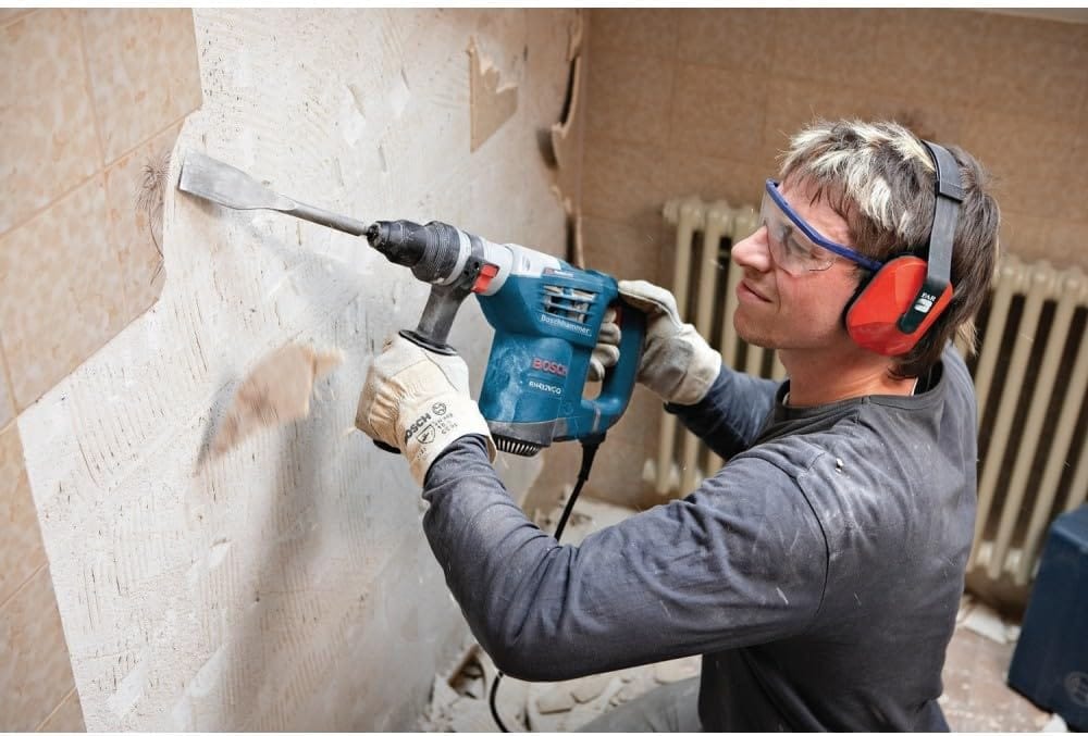 An application photograph of a renovation worker wearing clear safety glasses and red ear protectors while using the Bosch hammer drill fitted with a wide steel chisel bit to aggressively chip away old ceramic tiles from an interior bathroom wall.
