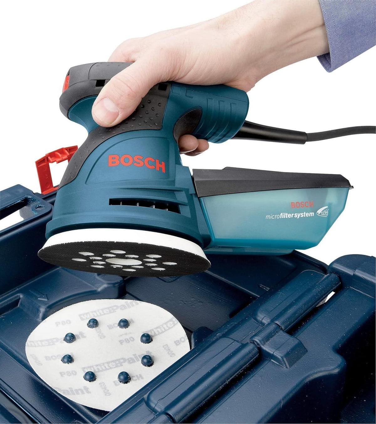 An application photograph of an operator holding the Bosch random orbit sander above its blue hard plastic carrying case, illustrating the 8-hole hook-and-loop pattern on the pliant white backing pad as it aligns with corresponding alignment pins on a circular sheet of sandpaper.