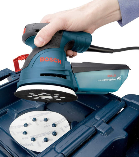 An application photograph of an operator holding the Bosch random orbit sander above its blue hard plastic carrying case, illustrating the 8-hole hook-and-loop pattern on the pliant white backing pad as it aligns with corresponding alignment pins on a circular sheet of sandpaper.