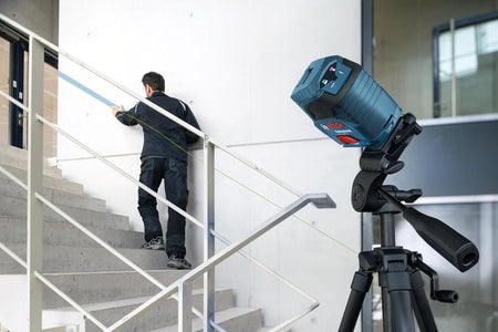 An action shot of the Bosch green beam laser mounted on a tripod at a steep angle, projecting a diagonal line for a professional installer to align a handrail along a concrete staircase.