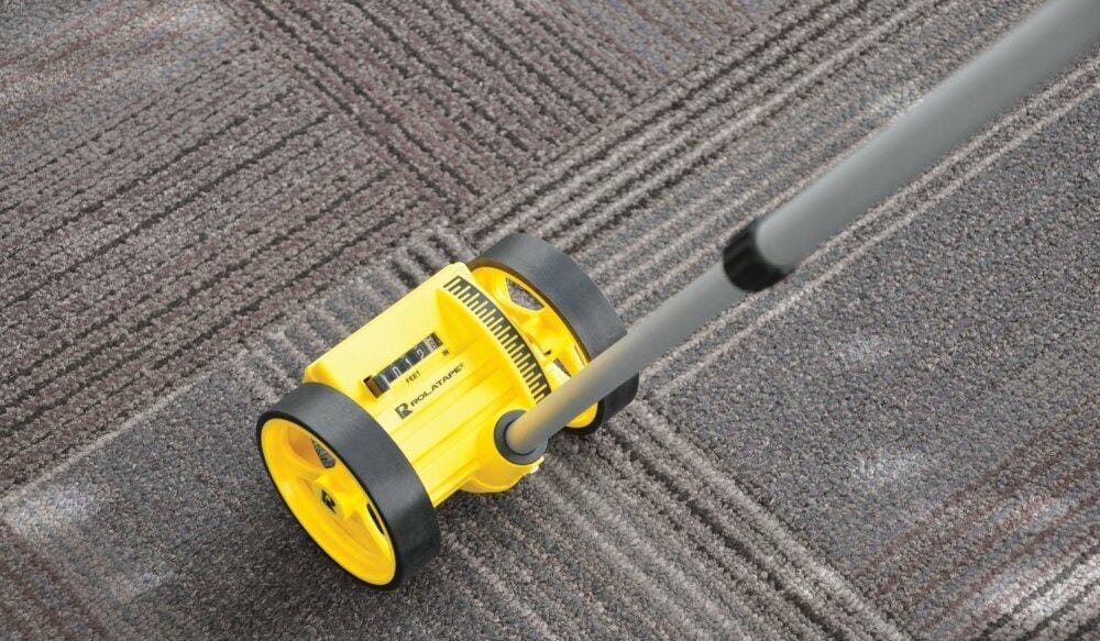 A close-up of the yellow Bosch Rolatape RT204 measuring wheel as it rolls across a patterned office carpet, with the counter visible.