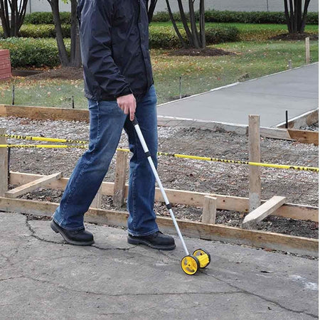 A person in jeans and a jacket uses the Bosch Rolatape RT204 measuring wheel to measure a paved walkway at an outdoor construction site.