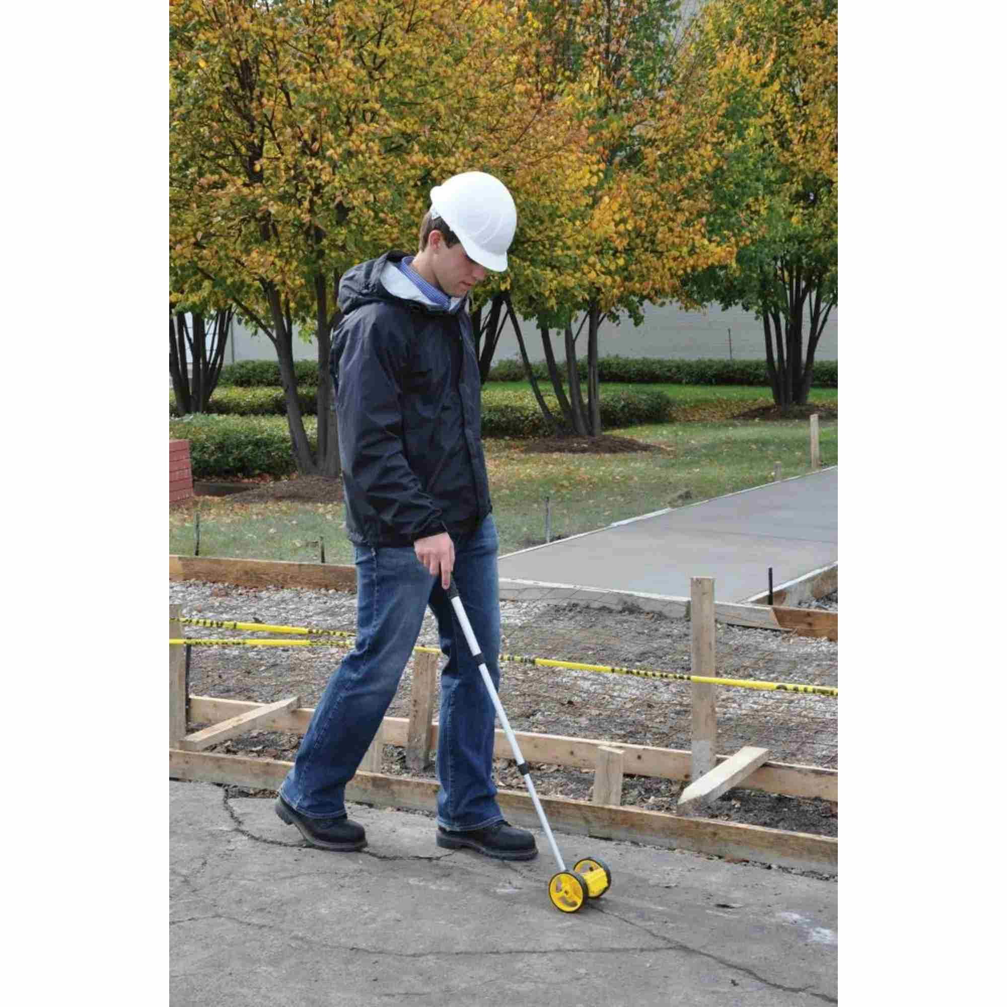 A person in a hard hat uses the Bosch Rolatape RT204 measuring wheel on a paved surface at a construction site next to concrete forms.