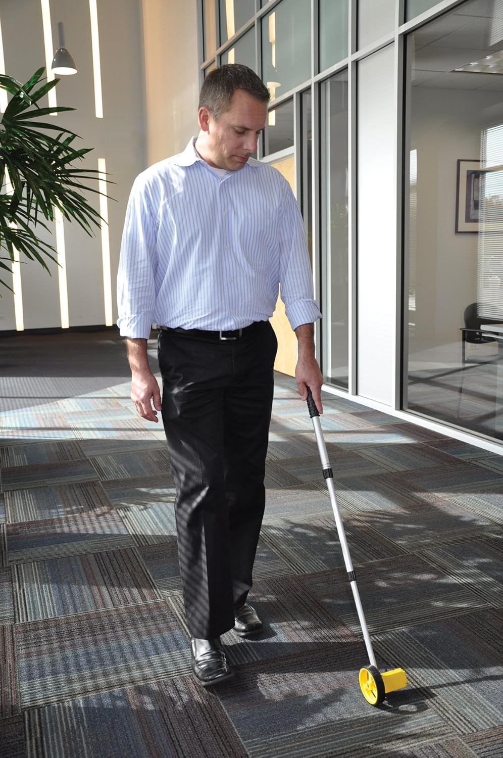 An indoor application lifestyle photo of a professional real estate agent or interior designer dressed in a striped dress shirt and black trousers, walking across a checkered commercial carpeted office floor while utilizing the yellow Bosch Rolatape RT104 4-inch single measuring wheel with its telescopic handle extended to map out space dimensions.