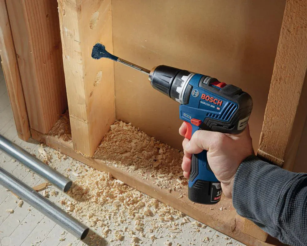 A real-world jobsite application photograph of a technician using a compact Bosch 12V cordless drill driver equipped with a blue spade paddle bit to bore a clean horizontal wire placement hole straight through a structural wood wall stud.