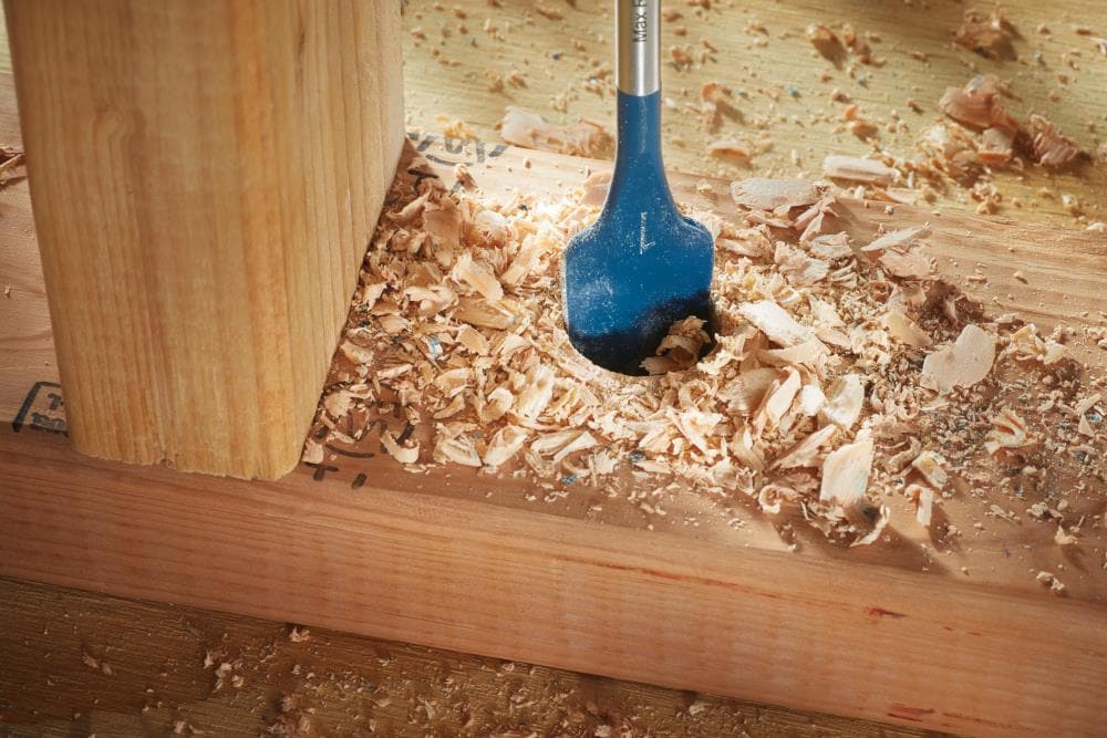 A macro close-up view of a blue Bosch woodboring spade bit finishing a large diameter hole in a piece of construction lumber, surrounded by heavy piles of freshly cleared wood shavings and chips.