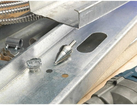 Action shot of a step drill bit resting on a galvanized steel channel next to a freshly drilled hole and conduit fitting, demonstrating electrical installation applications.