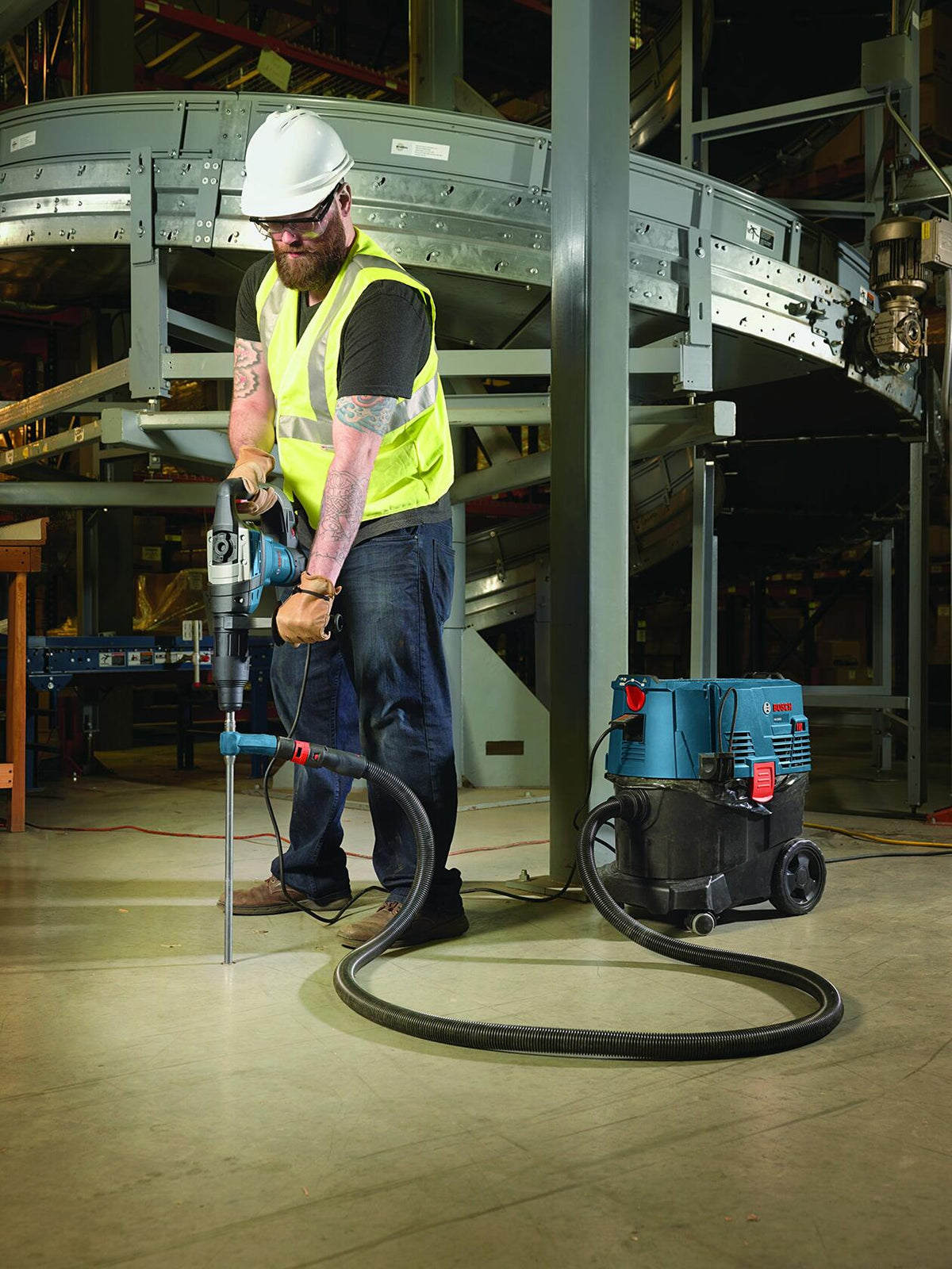 Worker in safety gear using Bosch drill with vacuum system to bore into concrete floor in industrial setting with conveyor system.