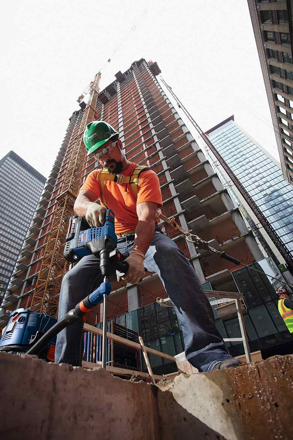 Low angle action shot looking up at a worker in an orange shirt drilling into a concrete column on a high-rise building construction site.