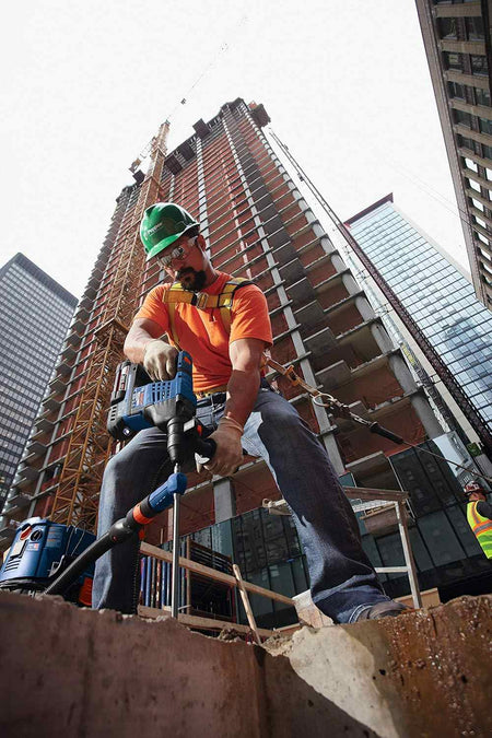 Low angle action shot looking up at a worker in an orange shirt drilling into a concrete column on a high-rise building construction site.