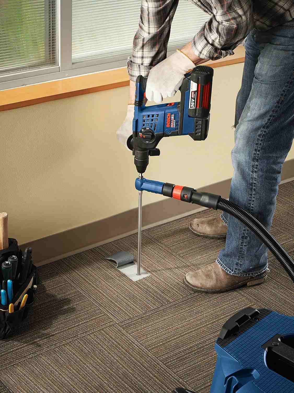 Action shot of a worker in a plaid shirt drilling vertically into a concrete floor through a carpet tile cutout in an office setting.