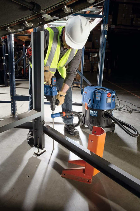 Action shot of a worker in a yellow vest drilling into a concrete floor under a conveyor belt system in an industrial facility.