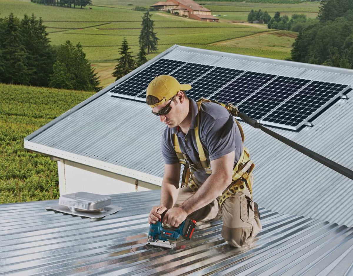 Action shot of a construction worker kneeling on a corrugated metal roof using a blue Bosch jigsaw to cut a ventilation hole.