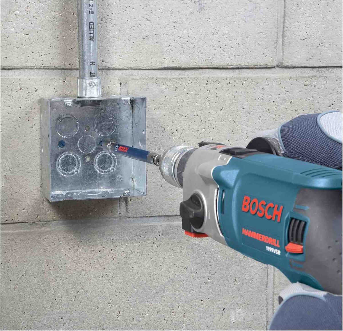 Close-up of a worker using a Bosch hammer drill equipped with a masonry bit to precisely drill a hole through a metal electrical box mounted on a concrete block wall.