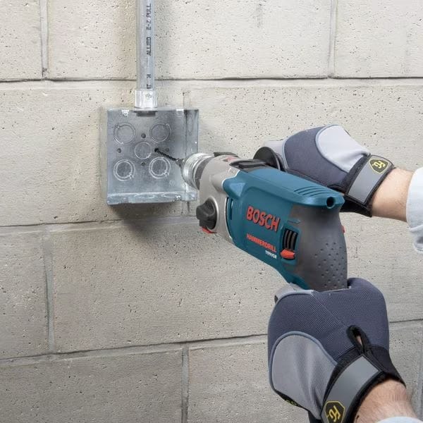 Worker wearing heavy-duty gloves operating a Bosch hammer drill with a masonry bit to secure a metal electrical junction box to a concrete block wall.