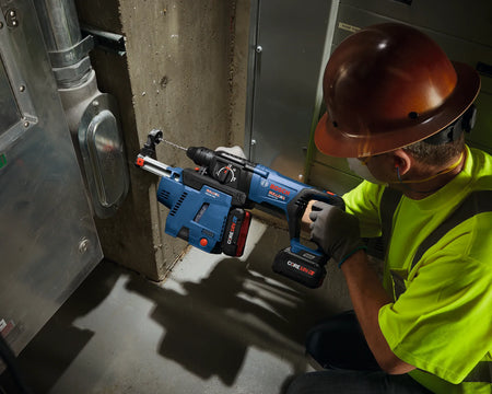 A professional commercial electrician kneeling inside a utility room to drill anchor holes into a solid reinforced concrete structural column. Equipped with a copper-tone hard hat, high-visibility vest, and safety glasses, the technician operates a heavy-duty Bosch 18V cordless rotary hammer drill outfitted with an integrated dust extraction vacuum attachment and an active carbide bit from the TCHC9 masonry set.