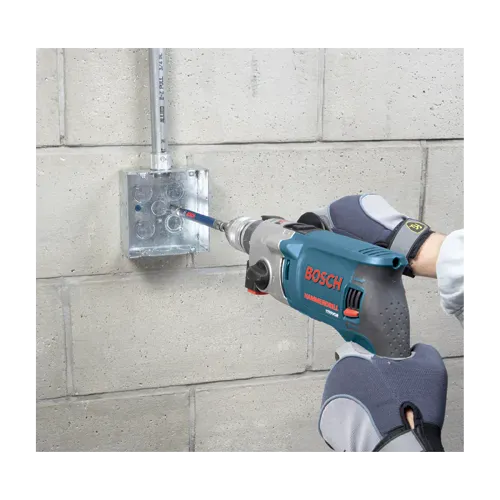 A wider field application shot identical to file TCHC9 7.webp showing an active trade specialist installing structural wiring conduit lines along a commercial masonry basement wall. The contractor utilizes both gloved hands to guide the blue Bosch hammer drill and matching steel sleeve guide attachment from the TCHC9 concrete screw kit to anchor the industrial electrical square box.