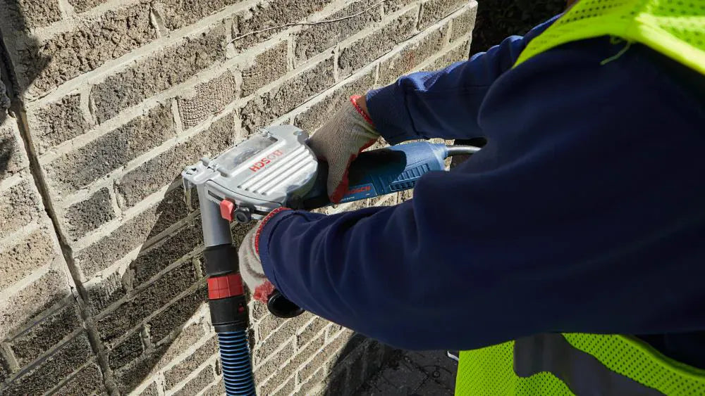 A contractor using a Bosch grinder equipped with the TG502 tuckpointing guard to remove mortar from a residential brick wall, demonstrating the effective dust capture with an attached vacuum.