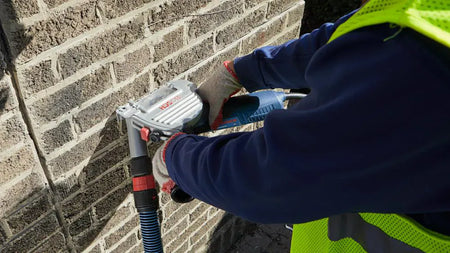 A contractor using a Bosch grinder equipped with the TG502 tuckpointing guard to remove mortar from a residential brick wall, demonstrating the effective dust capture with an attached vacuum.