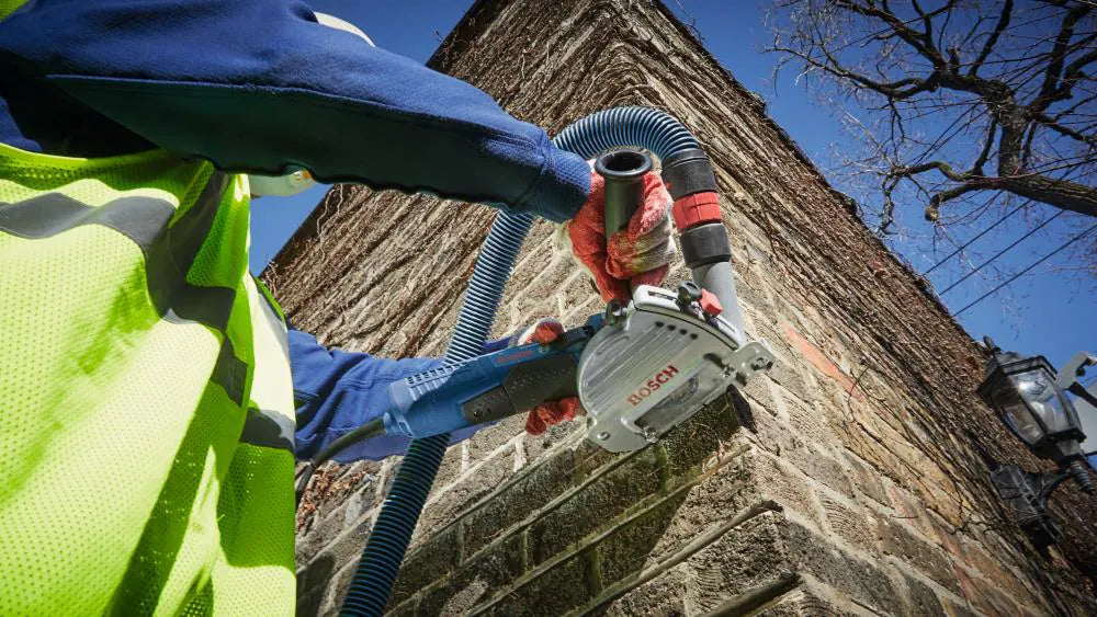 A low-angle action shot of the Bosch TG502 dust shroud in use on an exterior brick corner, emphasizing the tool's portability and the secure connection to the dust extraction system.