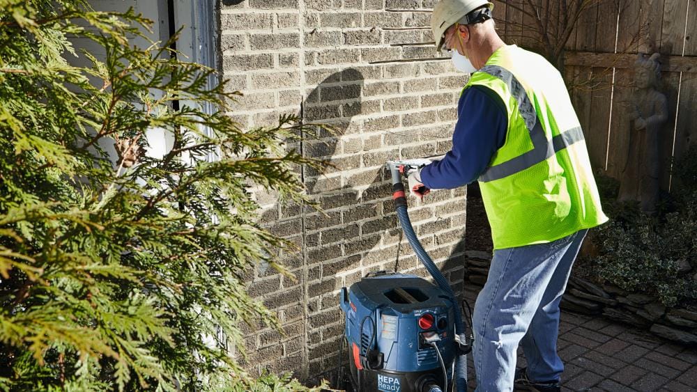 A wide shot of a masonry worker performing tuckpointing repairs with the Bosch TG502 guard and a HEPA-ready vacuum, illustrating a clean, OSHA-compliant workspace.