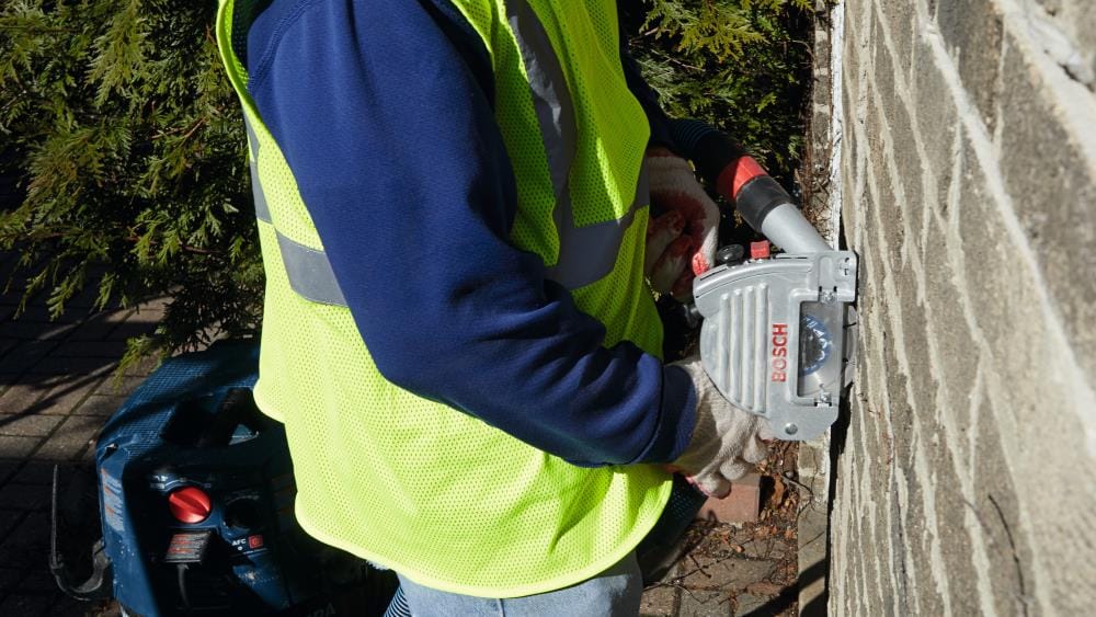 Profile view of the Bosch TG502 5-inch tuckpointing guard during vertical mortar removal, showing how the shroud stays flush against the brick for maximum dust containment.