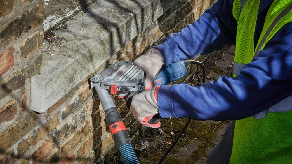 Masonry professional using the Bosch TG502 guard to clear mortar beneath a stone window sill, demonstrating the tool's versatility and compact design for tight spaces.