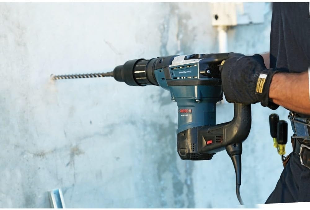 Worker using Bosch rotary hammer drill with long masonry bit to bore into a concrete wall, wearing gloves and tool belt in a construction setting.