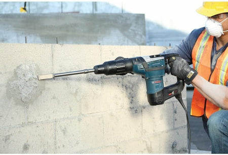 Person using a Bosch rotary hammer with chisel bit to break concrete block wall, wearing safety gear including gloves, mask, and high-visibility vest.